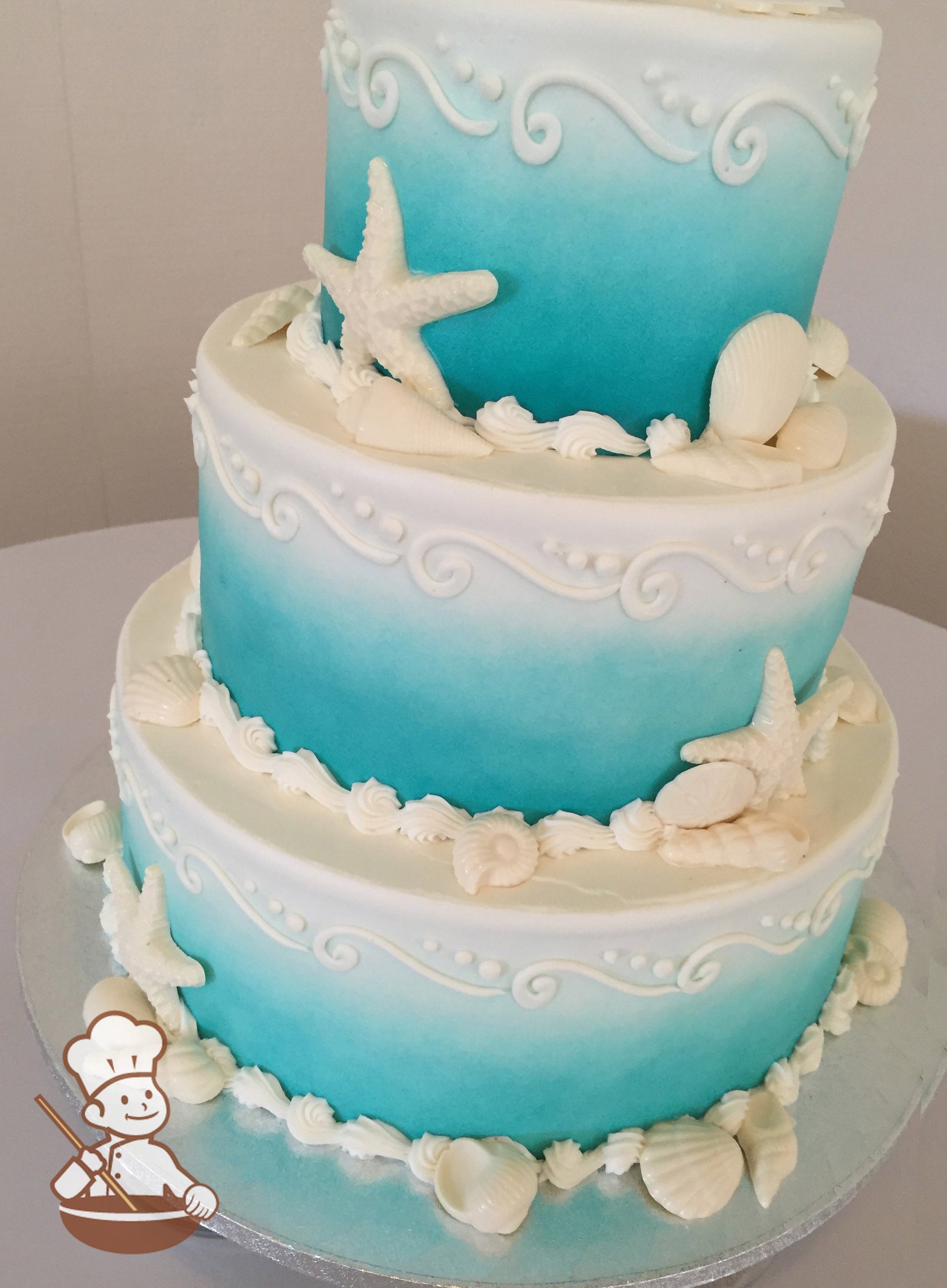 3-tier cake with smooth white icing and decorated with an ombre blue coloring, white buttercream scrolls and seashells.