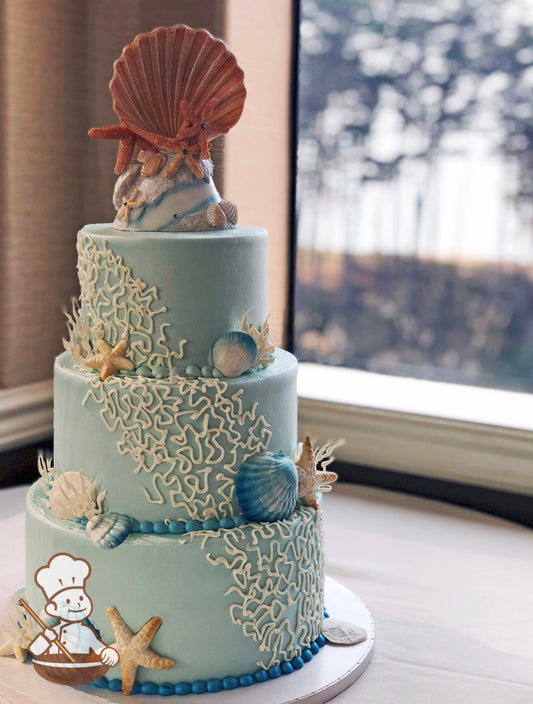 3-tier cake with light-blue smooth icing and decorated with a hand-piped white buttercream coral reef and fondant seashells.