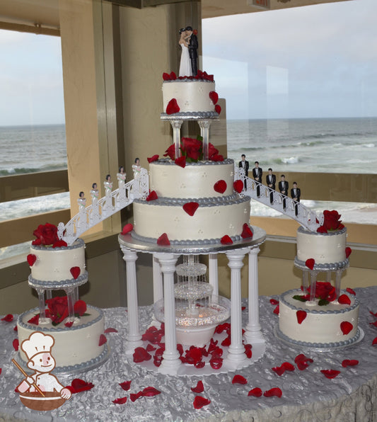 3 sets of cakes connected by two staircases with red rose petals. Center cake is elevated by plastic pillars surrounding a water fountain.