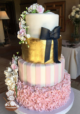 Her side cake decorated with sugar flowers, pink and white stripes, metallic gold, and black ribbon bow. Bottom tier in pinched pink ruffles.