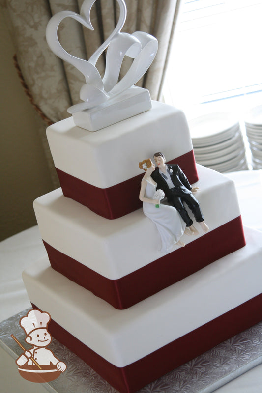 Square white fondant wedding cake with burgundy satin ribbon on base of each tier and decorated with double heart topper and sitting couple.